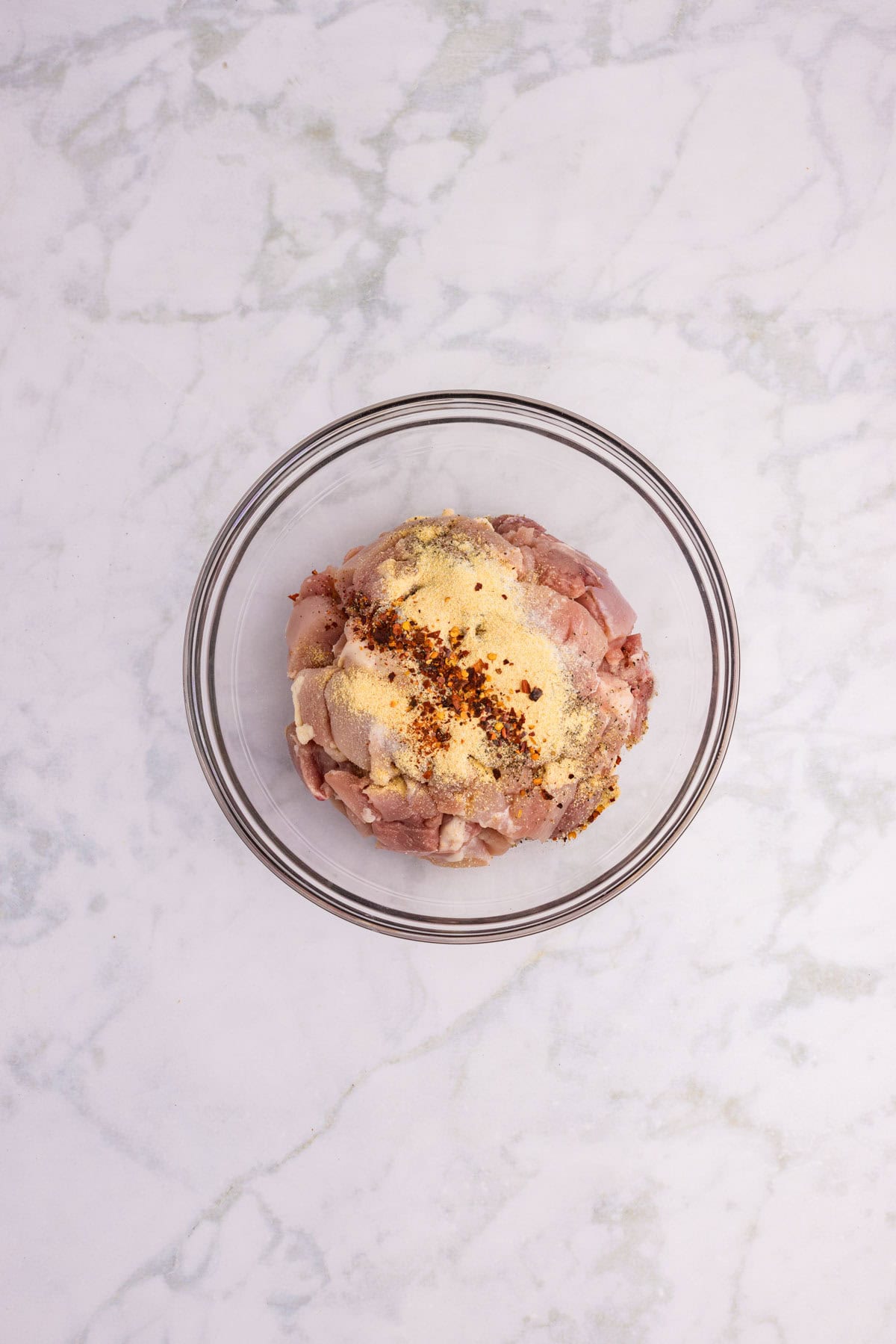 A glass bowl containing raw ground meat topped with spices, placed on a white marble surface.