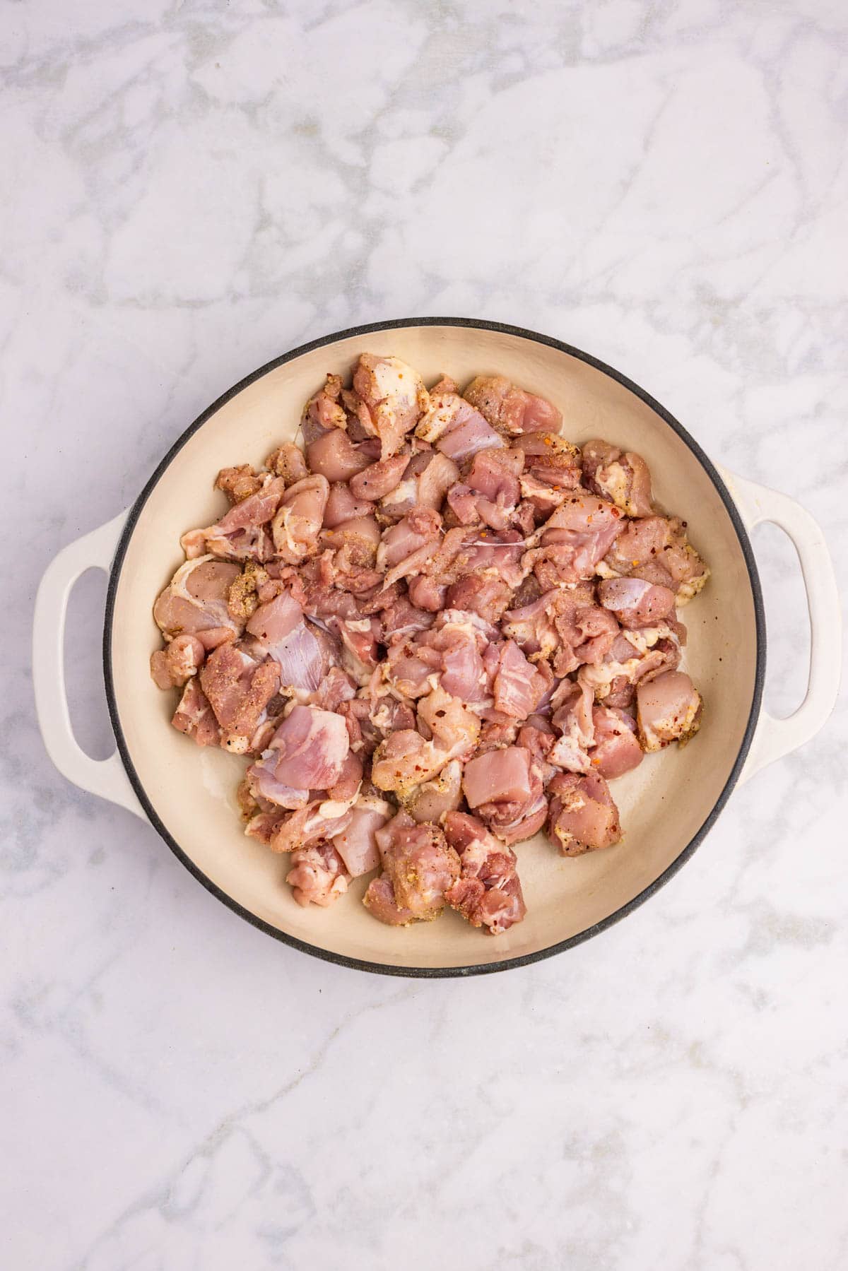 A round white dish with raw, seasoned pieces of chicken spread evenly inside, placed on a light marble surface.