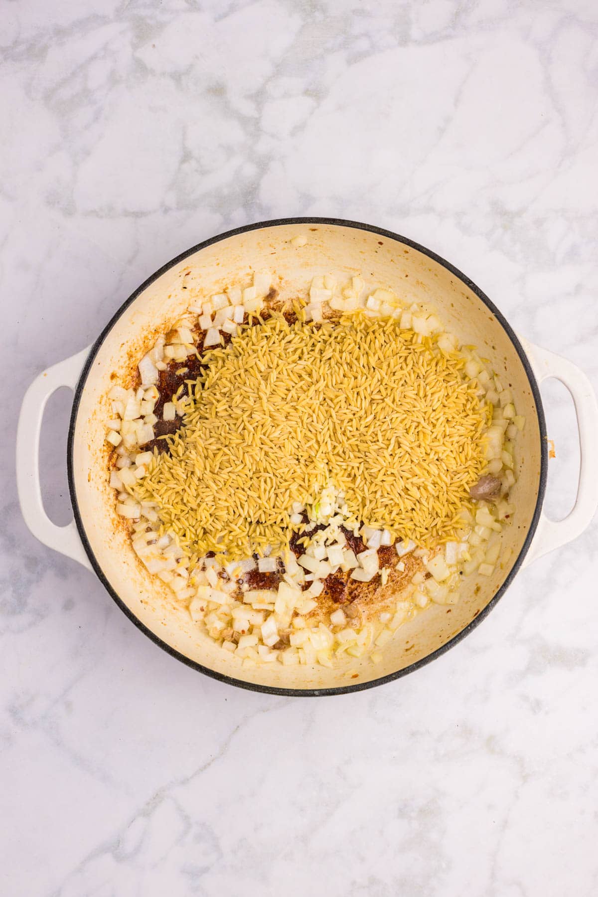 A white pot on a marble surface contains uncooked rice, chopped onions, and spices being saut&eacute;ed.