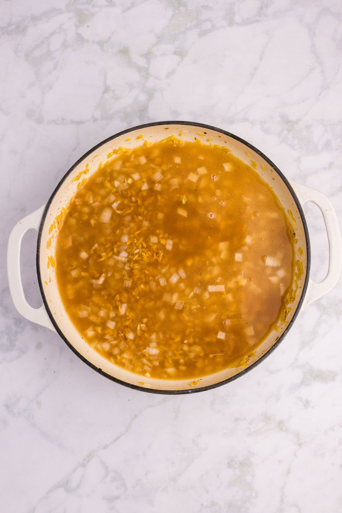 A white pot containing a mixture of chopped onions, rice, and broth sits on a marble countertop.