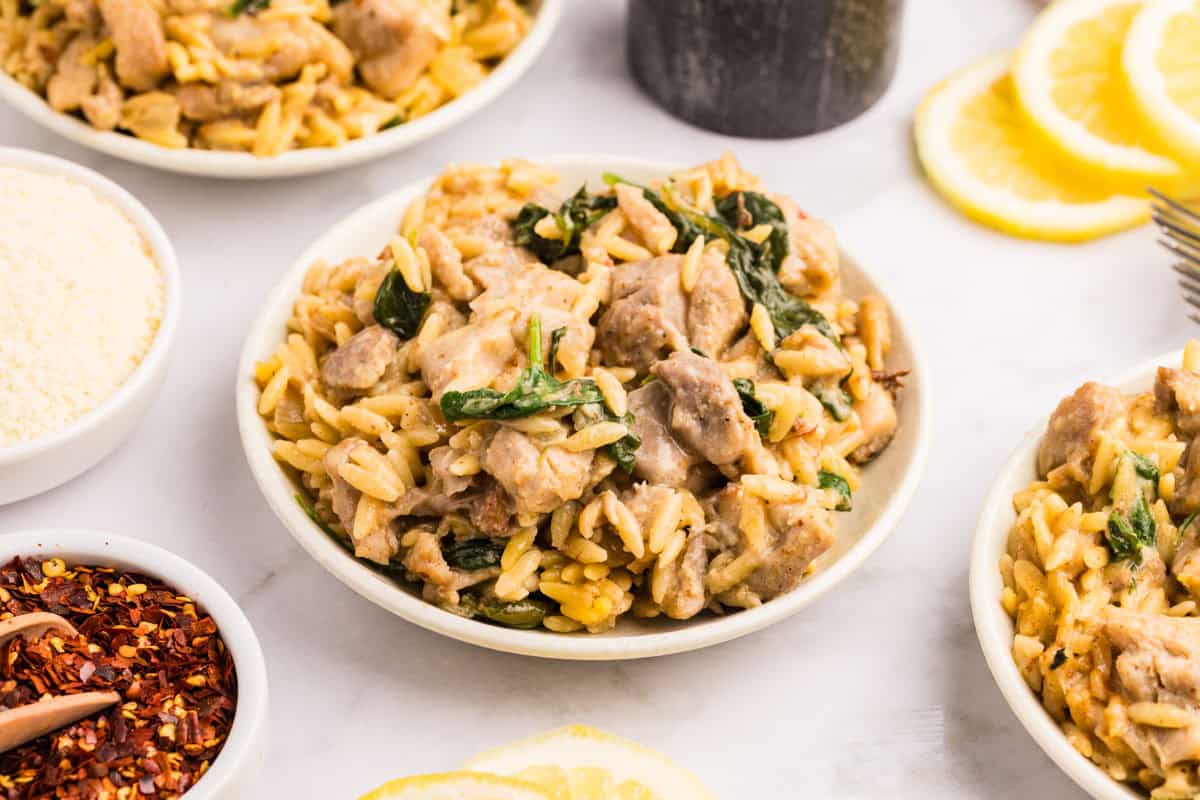 A plate of creamy orzo pasta with pieces of chicken and spinach, surrounded by small dishes of grated cheese and red pepper flakes, with lemon slices nearby.