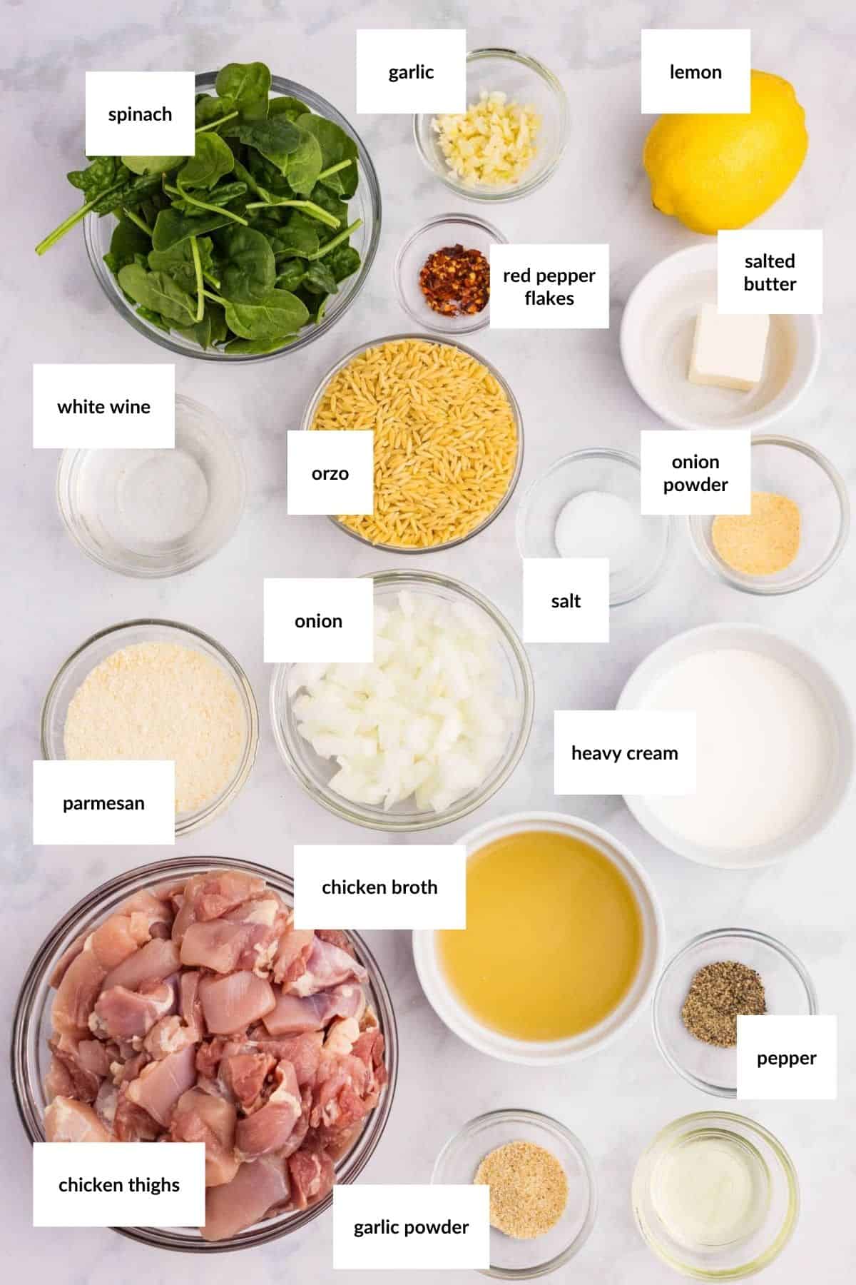 Various labeled ingredients for a chicken and orzo dish are arranged on a white surface, including chicken thighs, spinach, orzo, butter, seasonings, and dairy products.