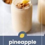 A glass of pineapple smoothie topped with granola and a pineapple slice, with a bowl of granola and pineapple pieces in the background. Text reads "pineapple smoothie.
