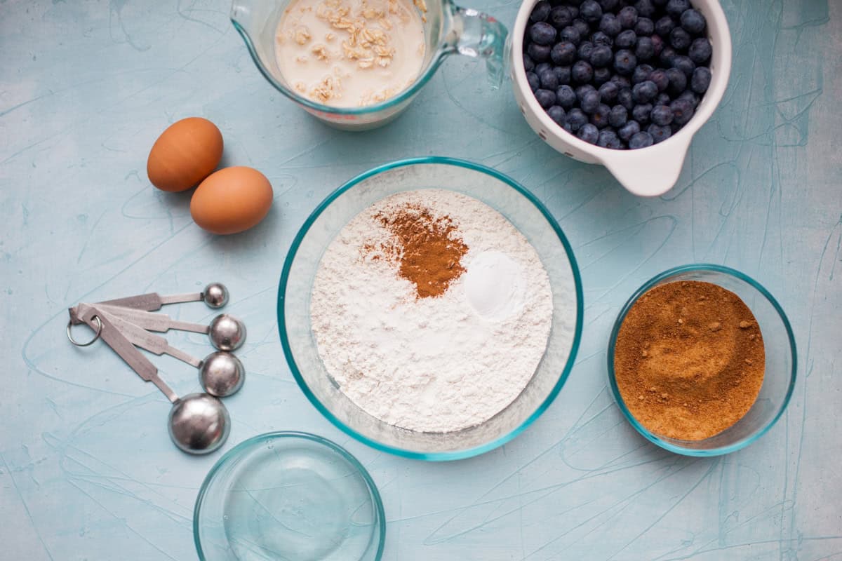Baking ingredients on a blue surface, including eggs, flour with spices, measuring spoons, oats in milk, blueberries, and brown sugar in bowls.