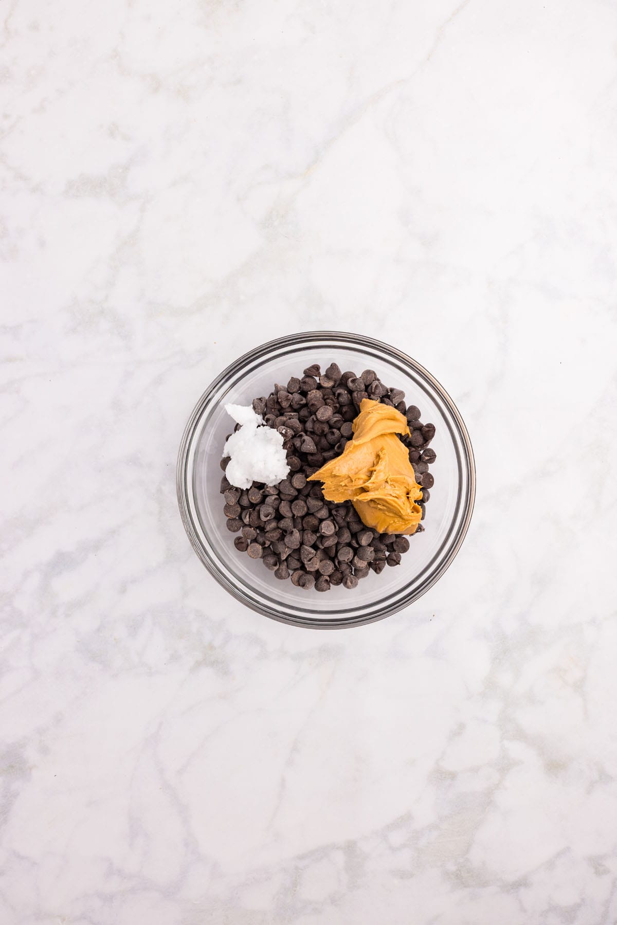 A glass bowl containing chocolate chips, a dollop of peanut butter, and a scoop of coconut oil on a white marble surface.