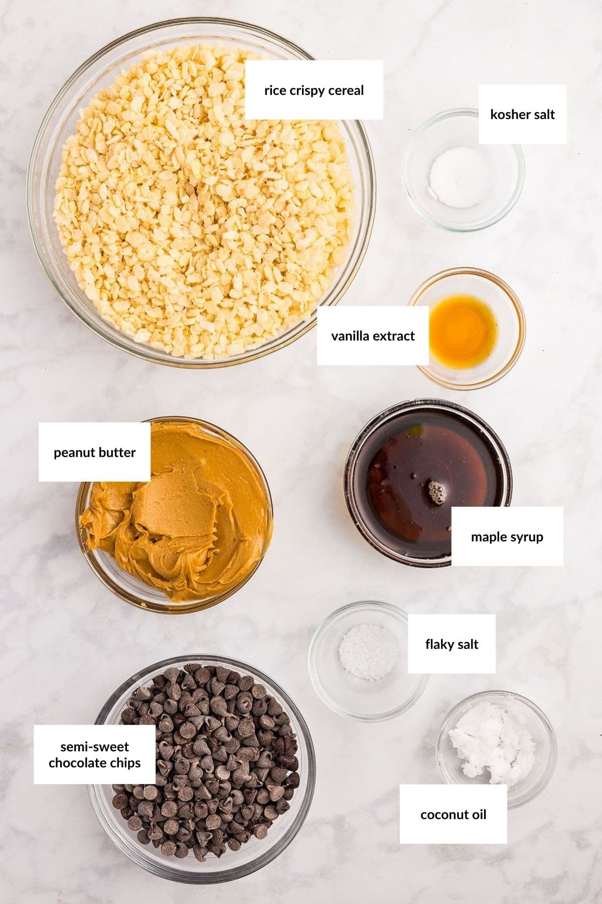 Top-down view of labeled ingredients in bowls on a marble surface: rice crispy cereal, kosher salt, vanilla extract, peanut butter, maple syrup, flaky salt, chocolate chips, and coconut oil.