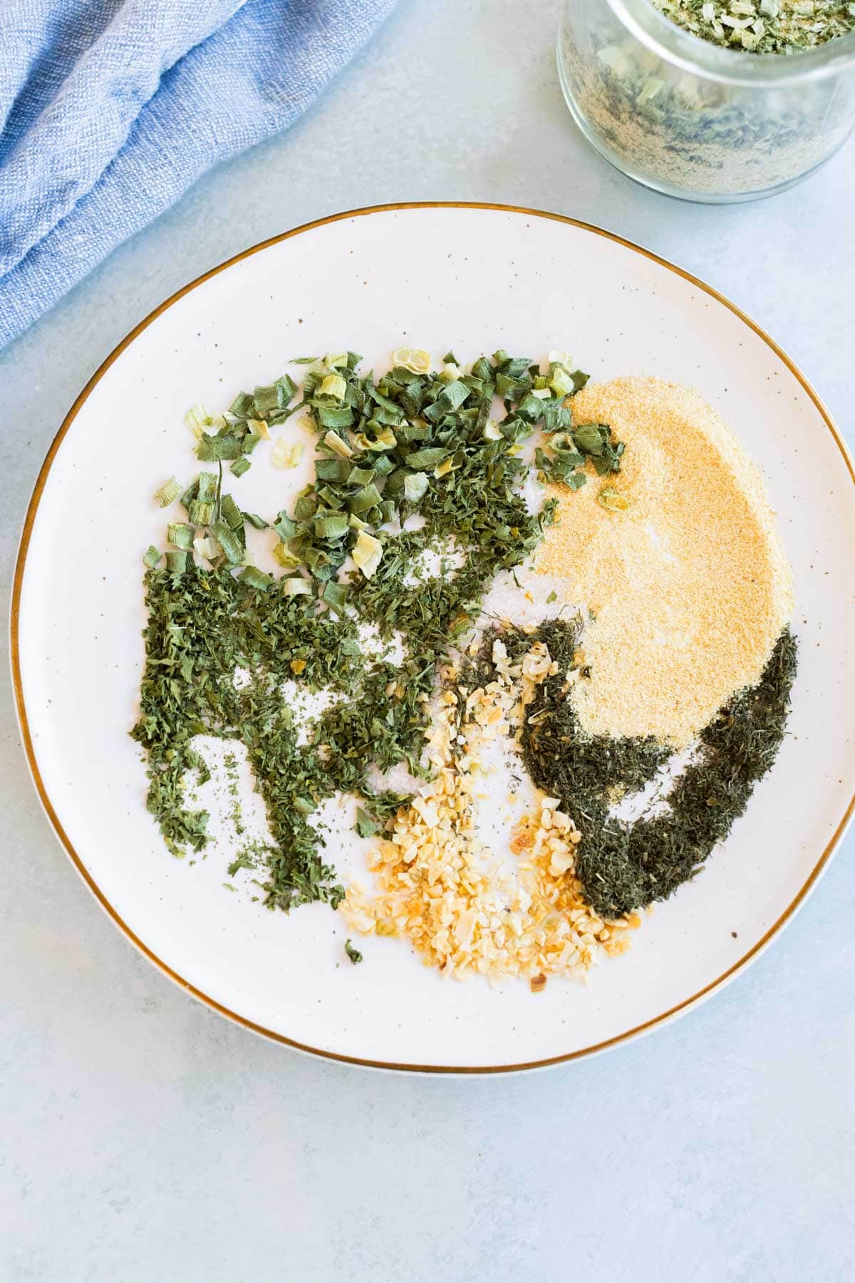 A white plate with separate piles of dried parsley, chives, minced garlic, onion powder, and dill, set on a light surface with a blue cloth and a jar of dried herbs nearby.