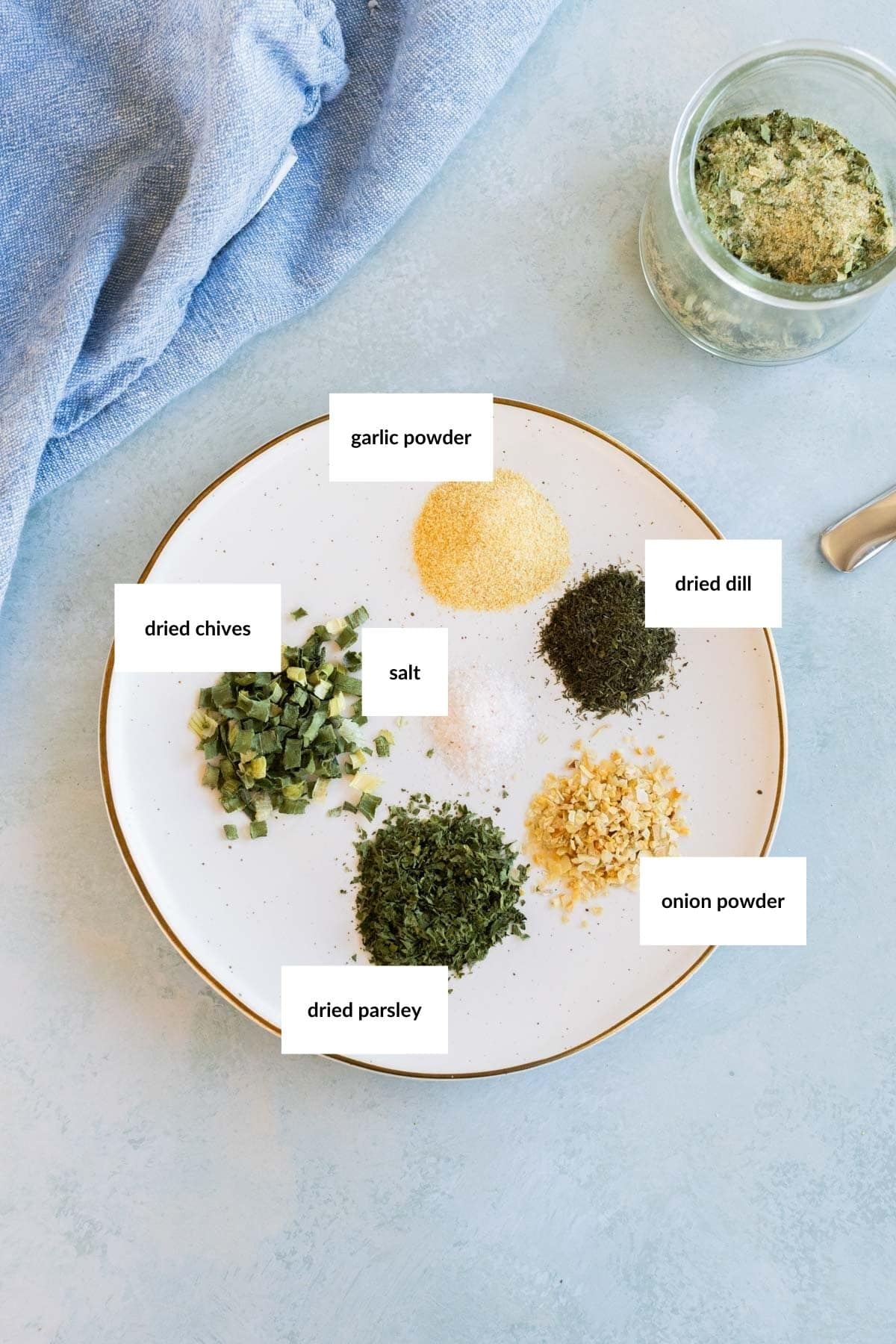 A white plate with labeled piles of garlic powder, dried dill, onion powder, dried parsley, dried chives, and salt, next to a jar of mixed herbs.