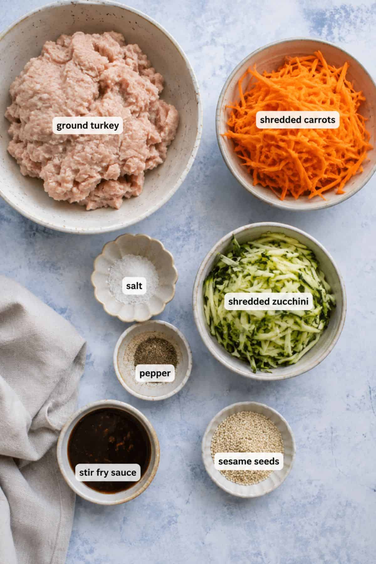 Bowls containing turkey stir fry made with ground turkey, shredded carrots, shredded zucchini, salt, pepper, stir fry sauce, and sesame seeds arranged on a light surface.