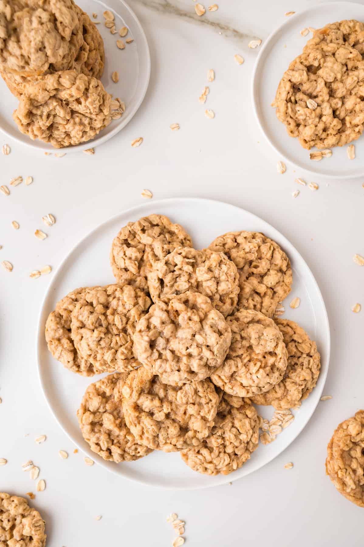A plate of oatmeal cookies arranged in a circle, with additional cookies on small plates and scattered oats on a white surface.