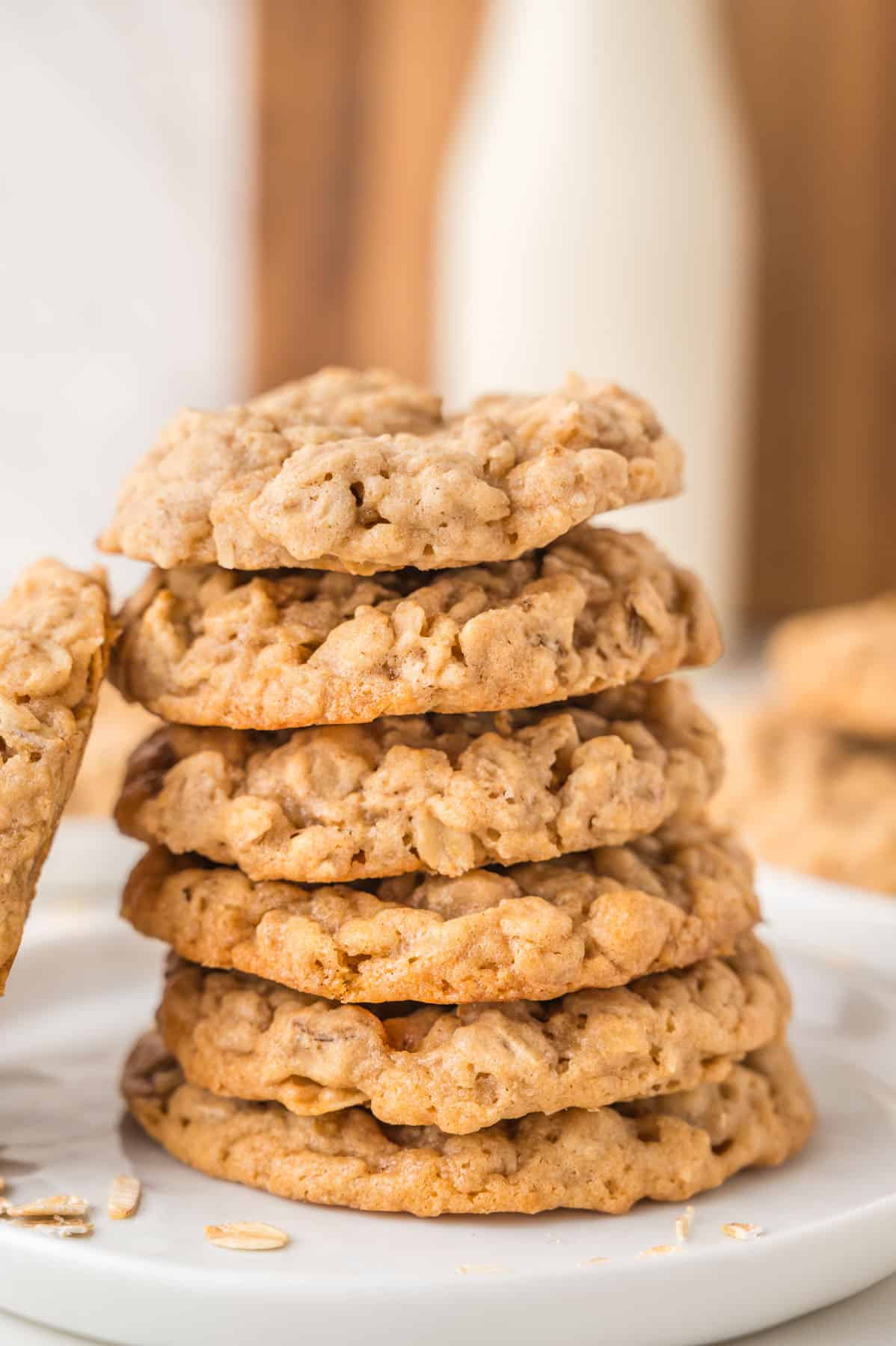 A stack of six oatmeal cookies sits on a white plate, with a bottle of milk and more cookies blurred in the background.