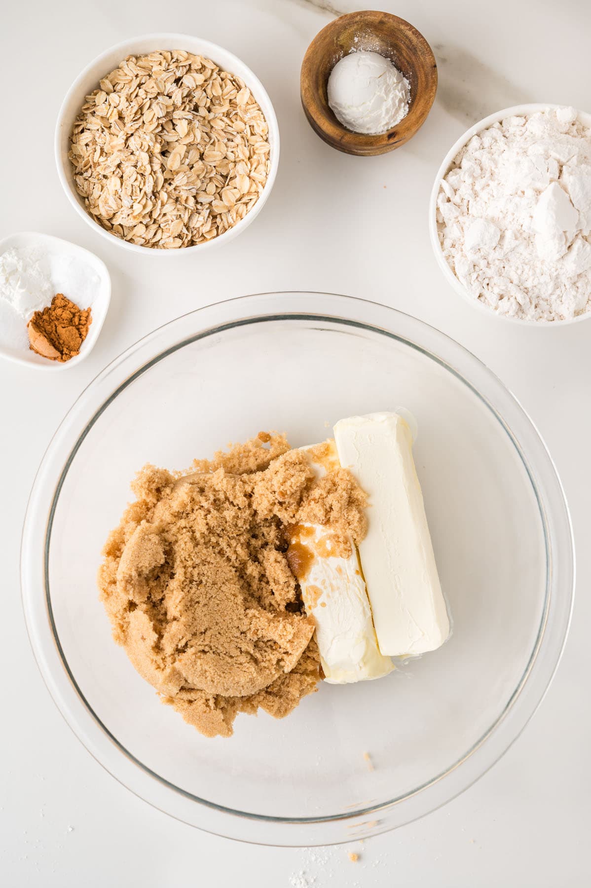 A glass bowl with brown sugar, butter, and vanilla, surrounded by small bowls of oats, flour, baking soda, and spices on a white surface.