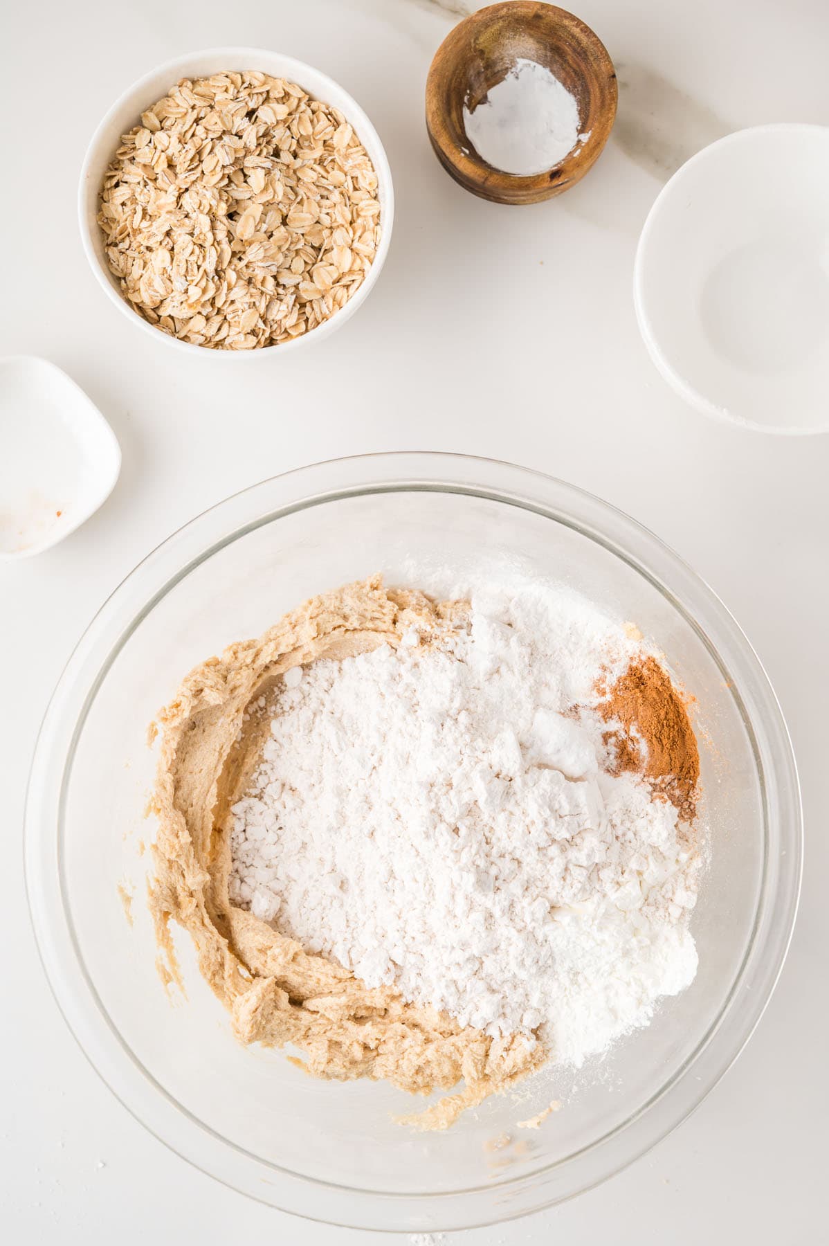 A glass mixing bowl with flour, cinnamon, and sugar on top of creamed butter, surrounded by bowls of oats, baking soda, and salt on a white surface.