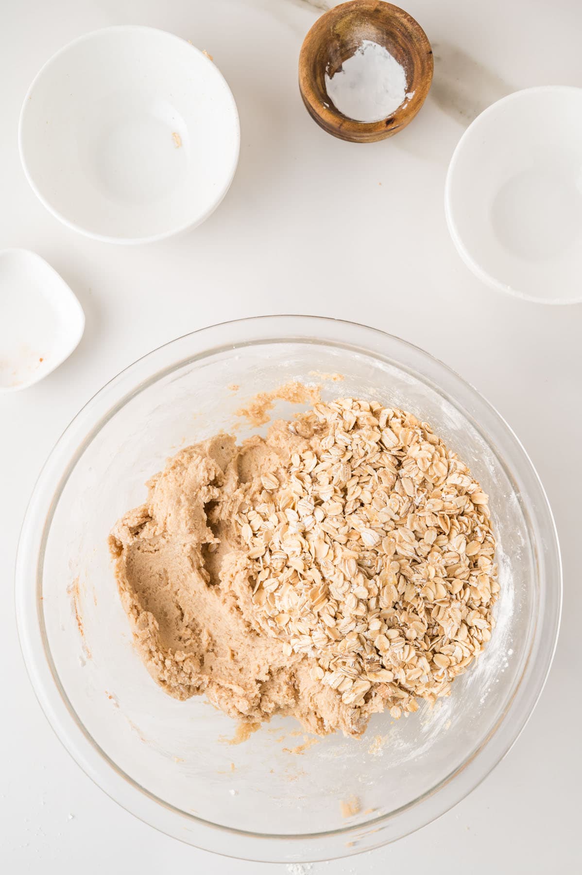 A glass bowl containing partially mixed cookie dough and oats sits on a white surface, surrounded by empty white dishes and a small wooden bowl with salt.
