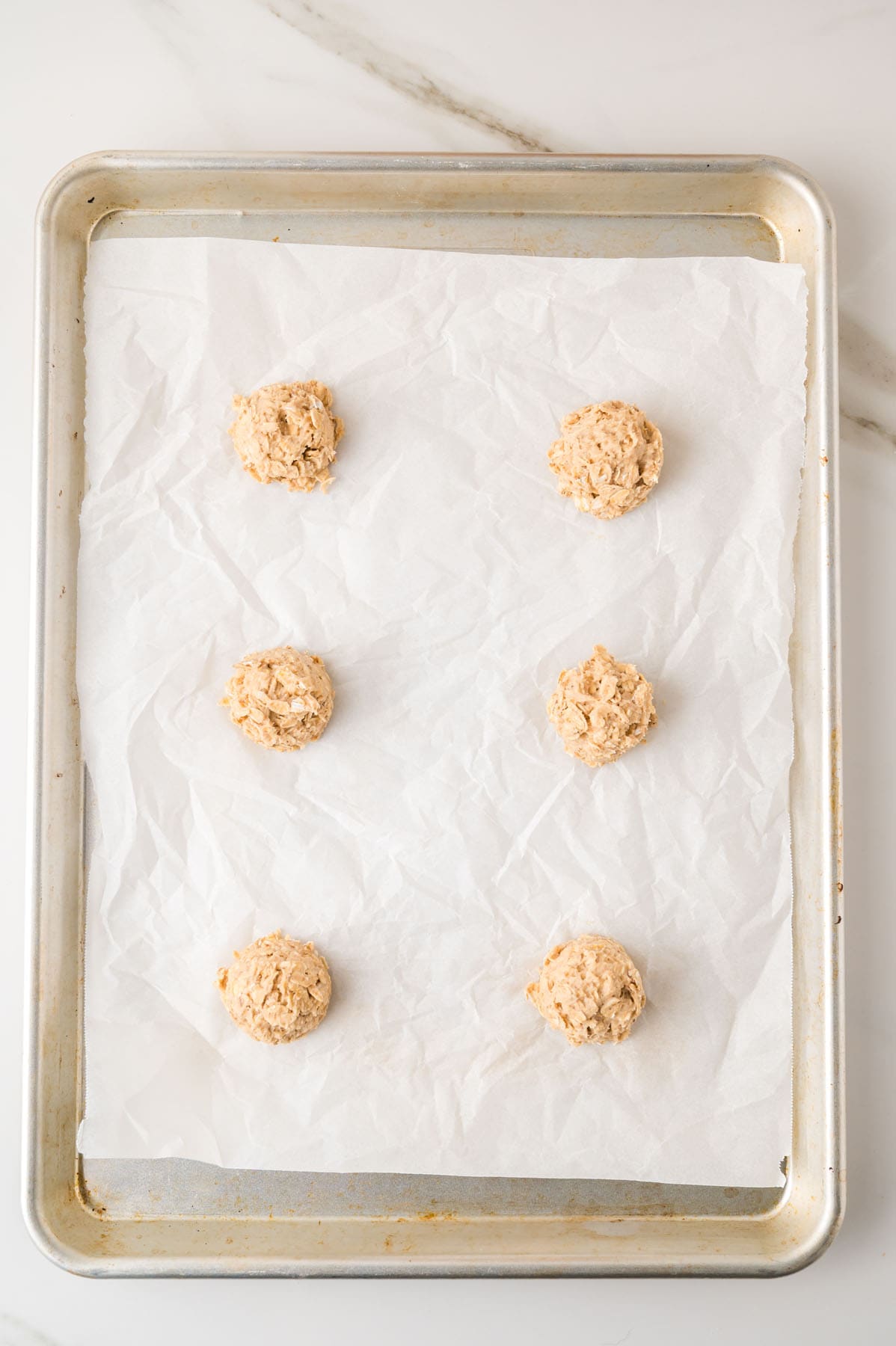 Six scoops of cookie dough are arranged on a parchment-lined baking sheet, ready to be baked.