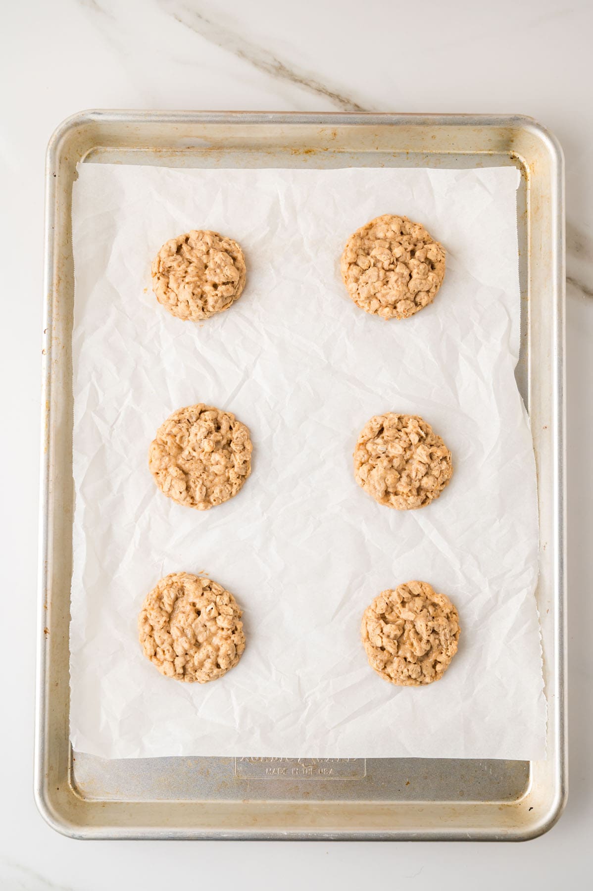 Six oatmeal cookies arranged in two columns on a parchment-lined baking sheet, viewed from above on a marble surface.