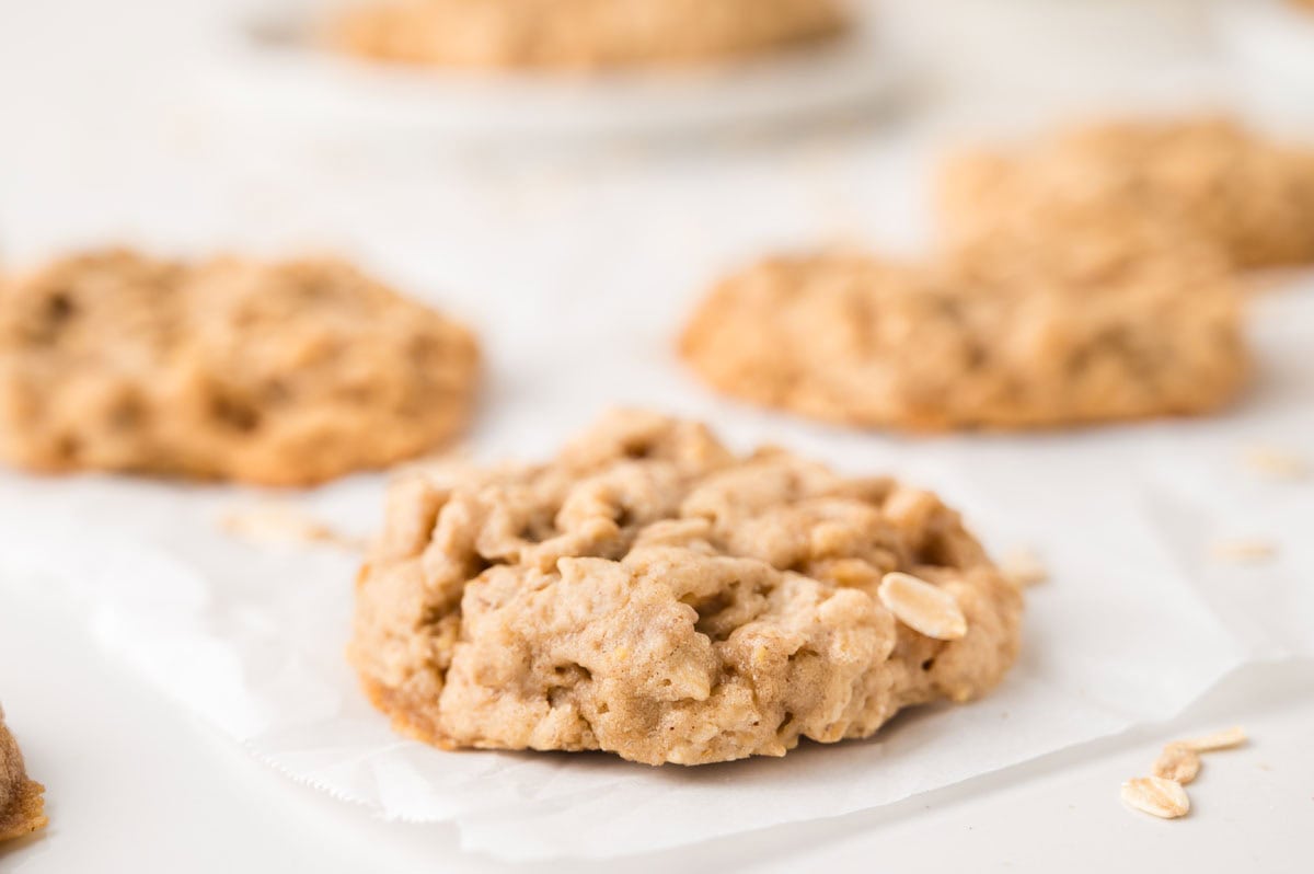 A close-up of an oatmeal cookie on parchment paper, with more cookies blurred in the background.