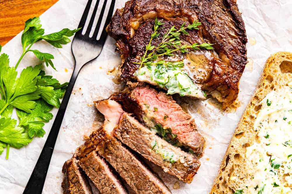 Sliced steak with herb butter and fresh herbs, served next to a slice of bread and a black fork on parchment paper.