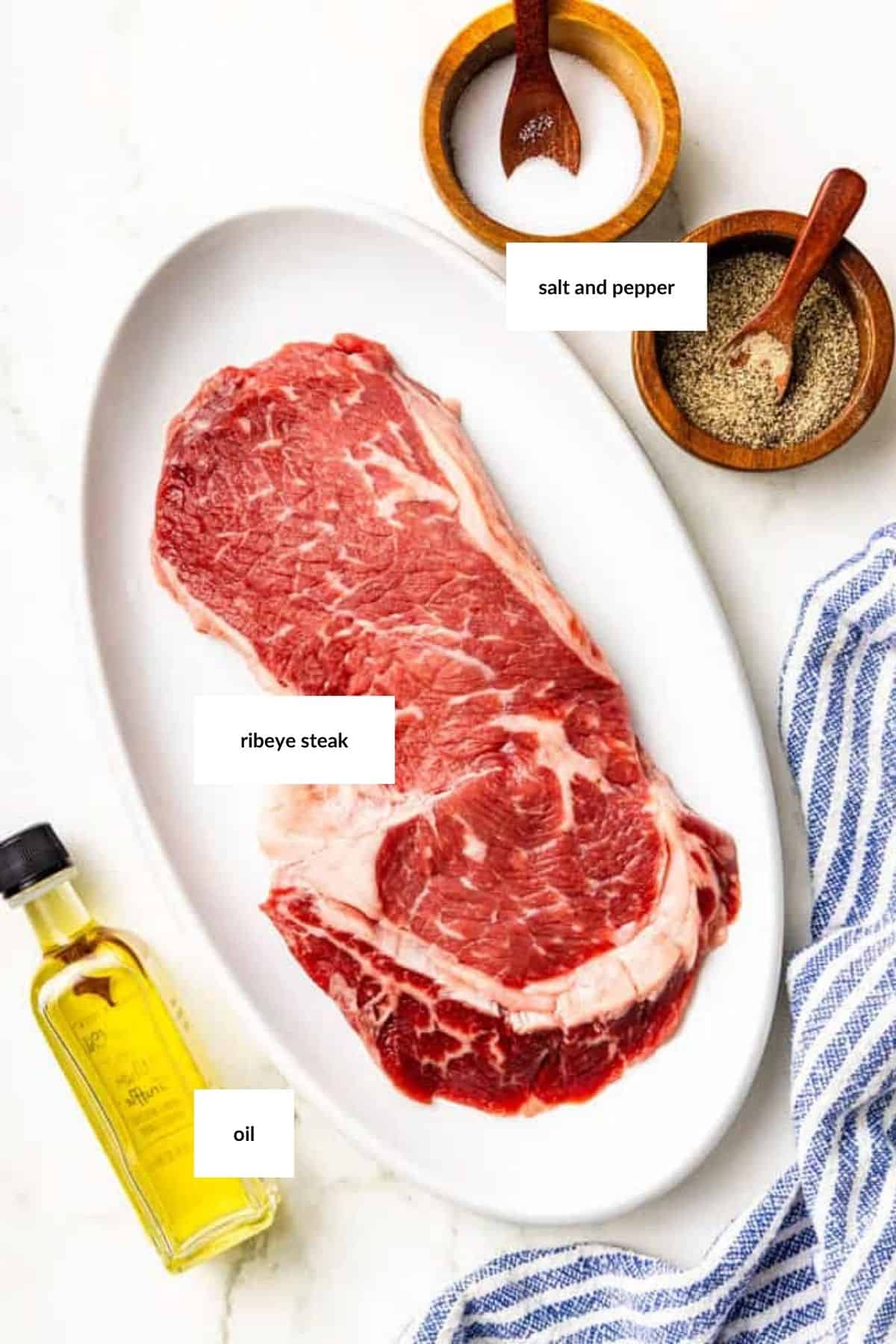 Raw ribeye steak on a white oval plate, with bowls of salt and pepper, a small bottle of oil, and a blue-striped towel beside it.