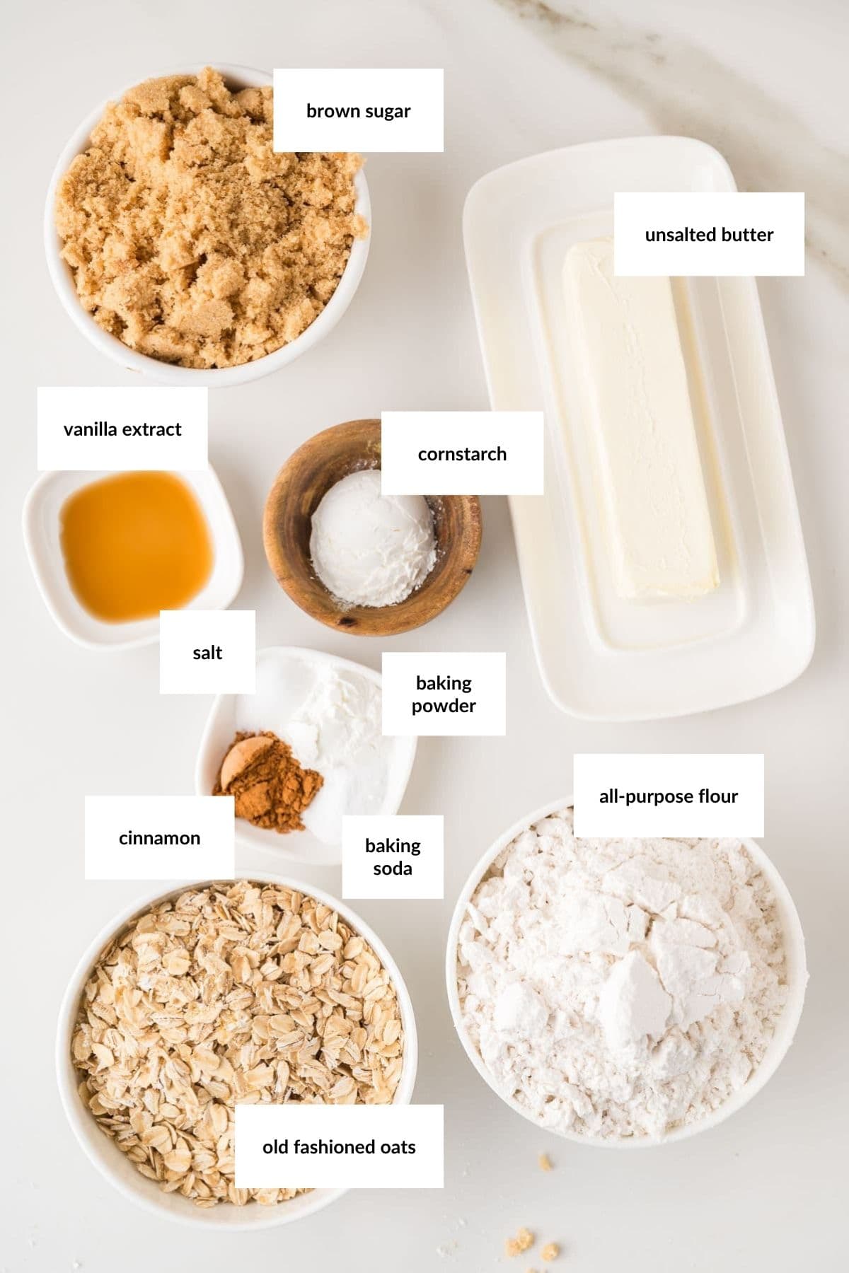 Bowls containing brown sugar, unsalted butter, vanilla extract, cornstarch, salt, baking powder, cinnamon, baking soda, all-purpose flour, and old fashioned oats on a white surface.