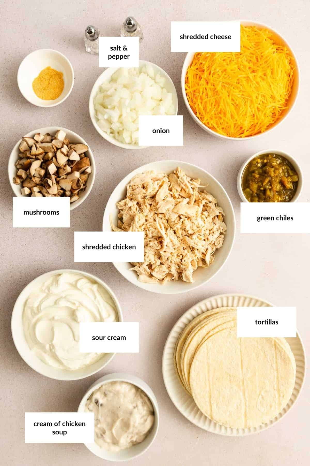 Top-down view of labeled ingredients in bowls and plates: shredded cheese, onion, green chiles, mushrooms, shredded chicken, sour cream, cream of chicken soup, tortillas, salt, and pepper.