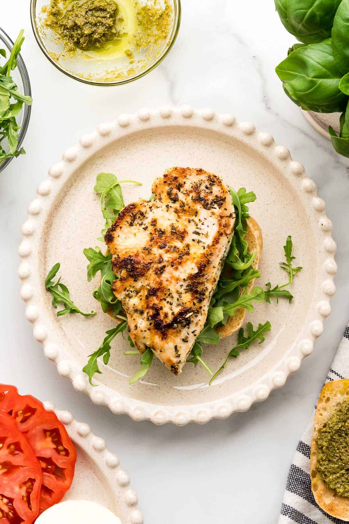 Grilled seasoned chicken breast on a bed of arugula atop a toasted bun, with pesto, tomato slices, mozzarella, and fresh basil nearby.