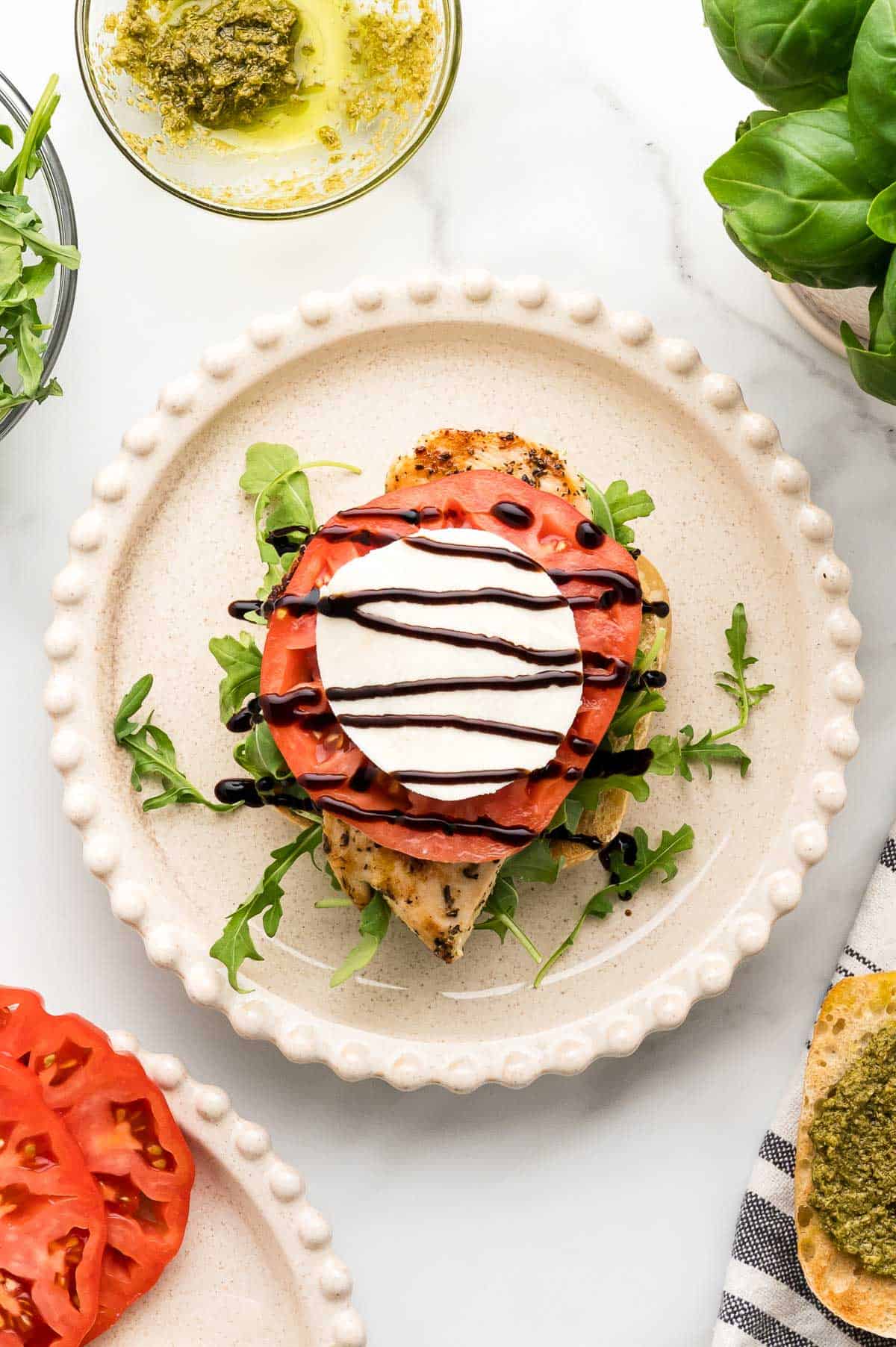 A plate with grilled chicken breast topped with arugula, tomato slices, mozzarella, and drizzled with balsamic glaze. A bowl of pesto and fresh basil are nearby.