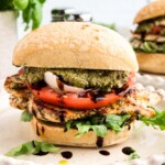 Grilled chicken sandwich with arugula, tomato, mozzarella, pesto, and balsamic glaze on a ciabatta bun, served on a white plate.