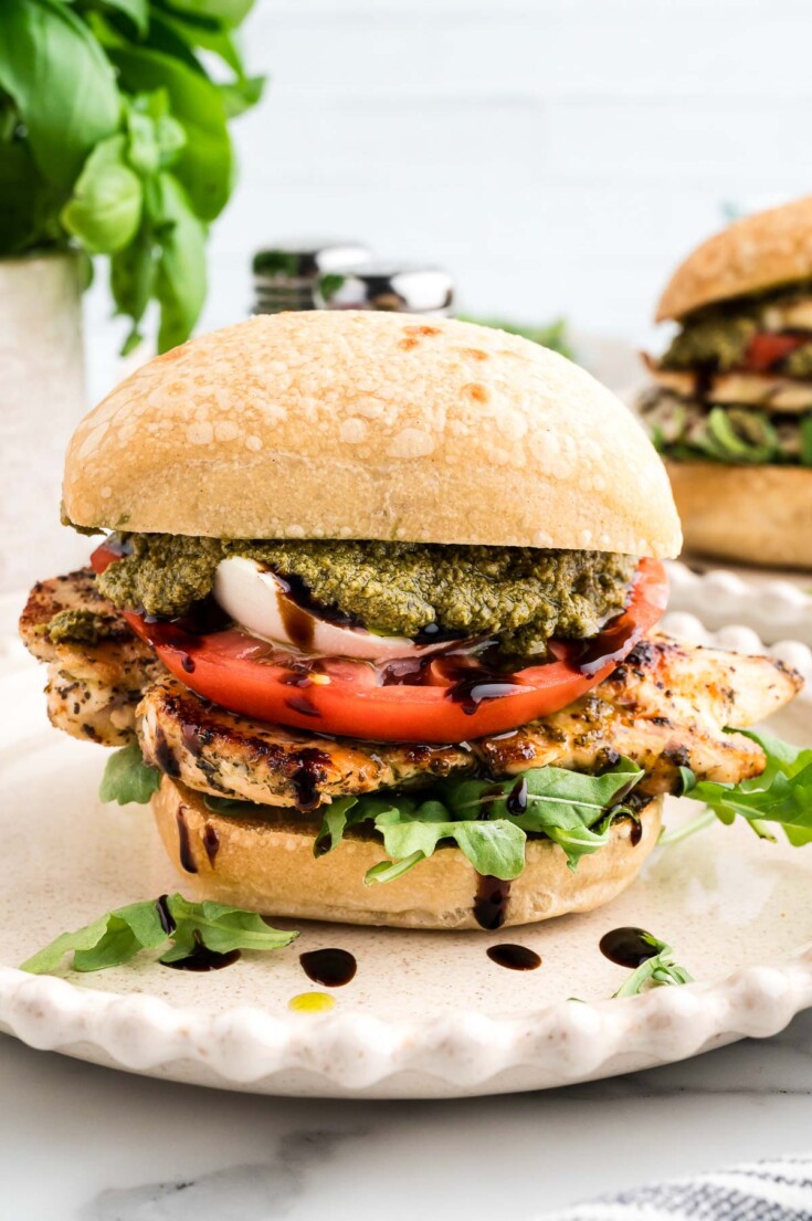 Grilled chicken sandwich with arugula, tomato, mozzarella, pesto, and balsamic glaze on a ciabatta bun, served on a white plate.