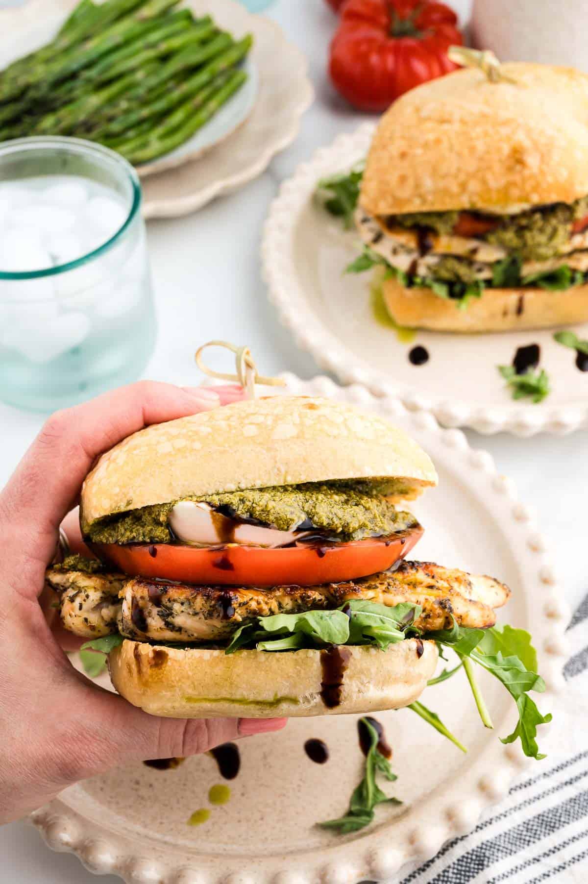 A hand holds a grilled chicken sandwich with tomato, arugula, pesto, and balsamic glaze on ciabatta bread. Another sandwich, asparagus, and a glass of ice water are on the table.
