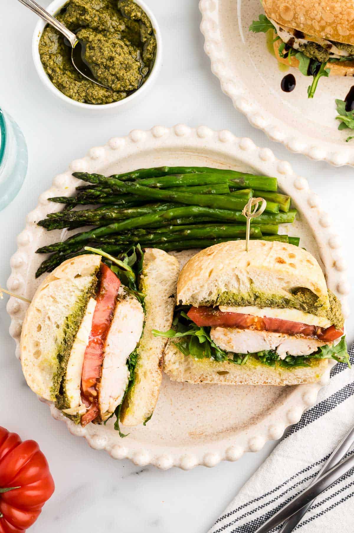 A sliced chicken sandwich with pesto, lettuce, and tomato on a plate, served with a side of cooked asparagus spears.