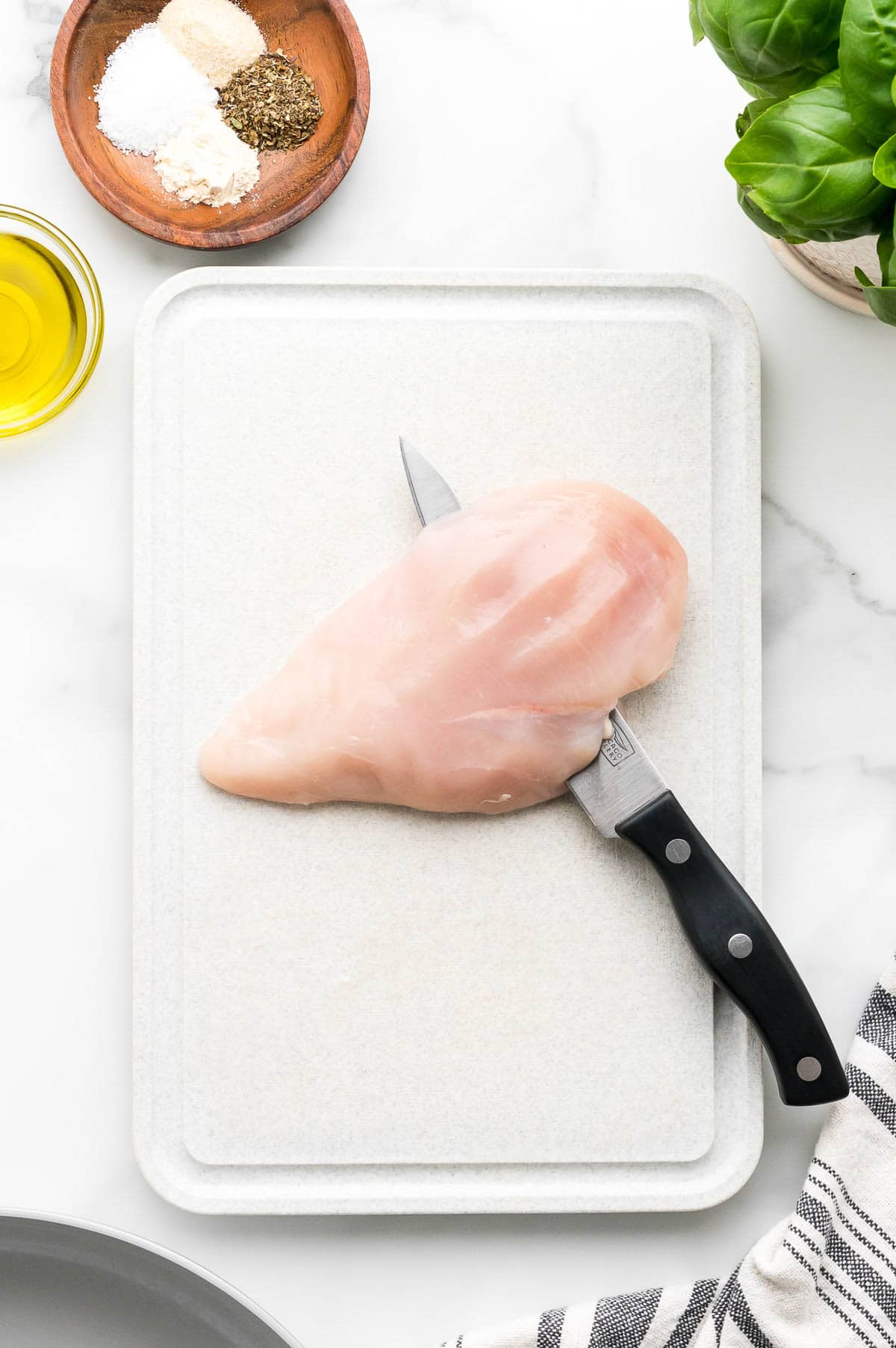 Raw chicken breast on a cutting board with a knife, surrounded by olive oil, spices in a wooden dish, and fresh basil leaves.