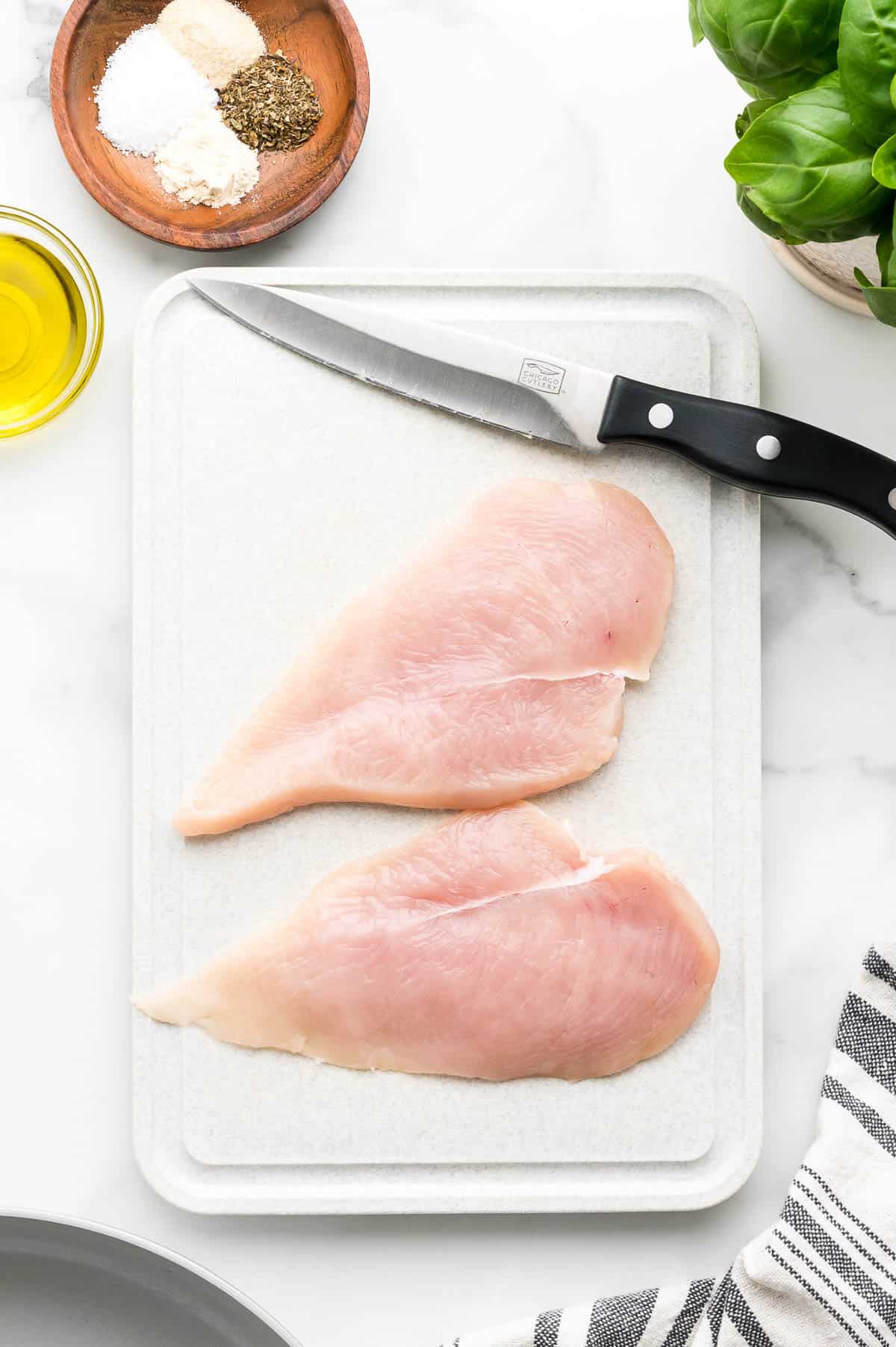 Two raw chicken breasts on a white cutting board with a knife, next to a bowl of spices, a small dish of olive oil, and fresh basil.