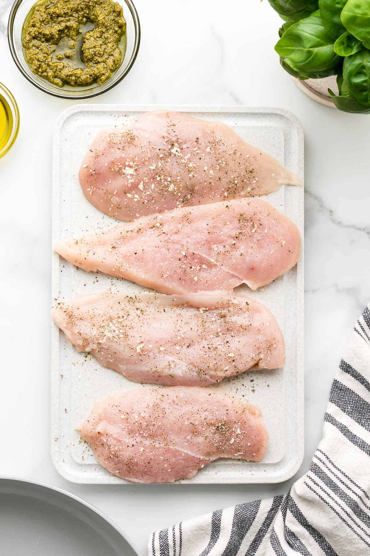Four raw, seasoned chicken breasts are arranged on a white cutting board. A bowl of pesto, fresh basil, and a striped towel are nearby.