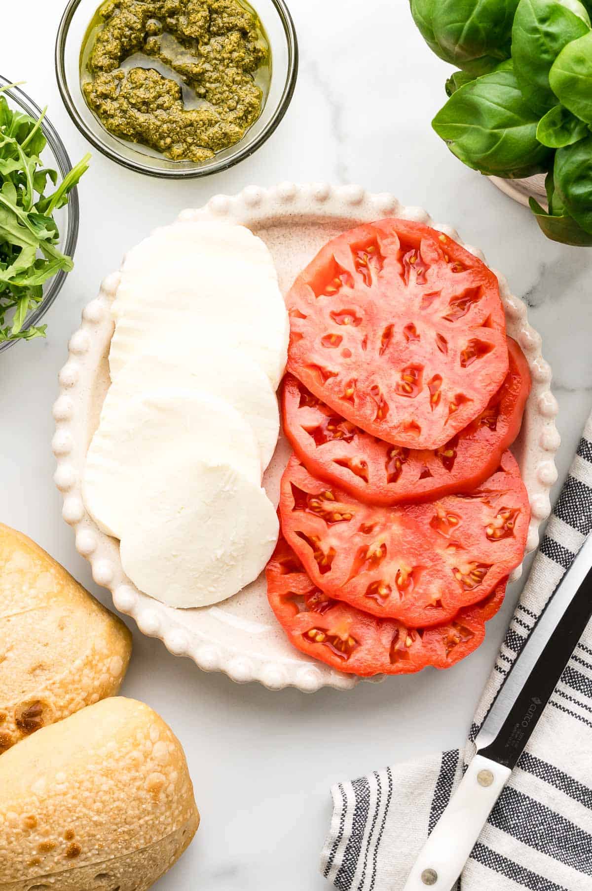 A plate with sliced mozzarella cheese and tomato, surrounded by fresh basil, arugula, ciabatta bread, pesto, and a knife on a striped towel.