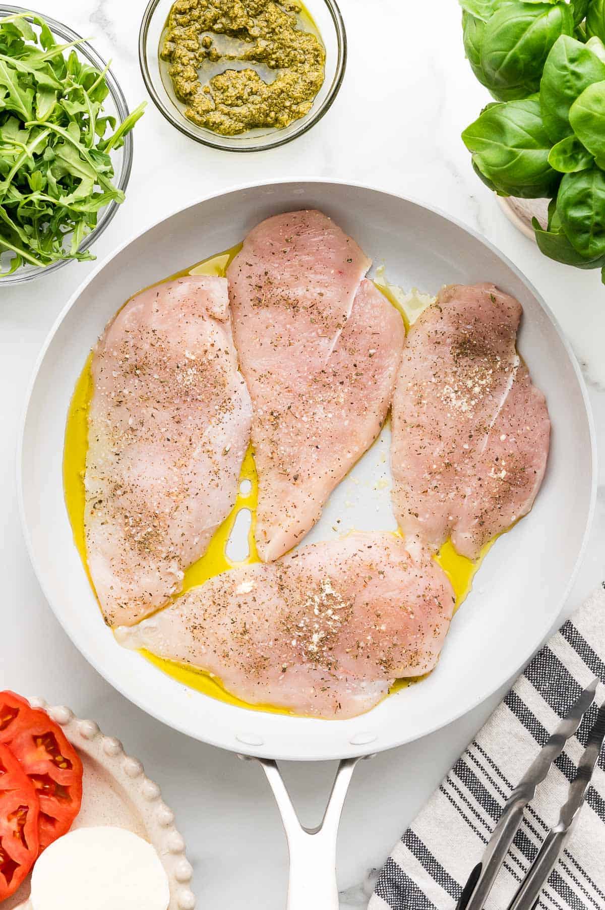 Raw chicken breasts seasoned with pepper and oil in a white skillet, surrounded by bowls of pesto, arugula, fresh basil, tomato slices, and mozzarella.