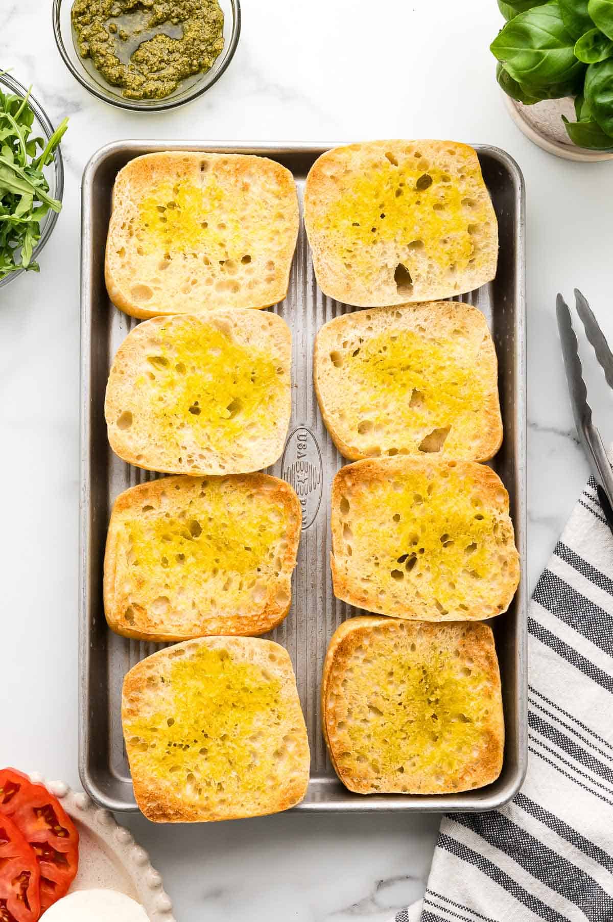Eight slices of toasted ciabatta bread with olive oil are arranged on a baking sheet; pesto, arugula, basil, and tomato are visible nearby.