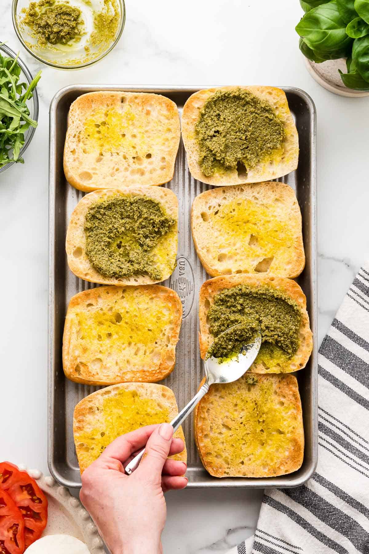 A hand spreads green pesto onto toasted ciabatta buns on a baking sheet; some buns have olive oil, while others have pesto. Nearby are a bowl of pesto, arugula, and basil.