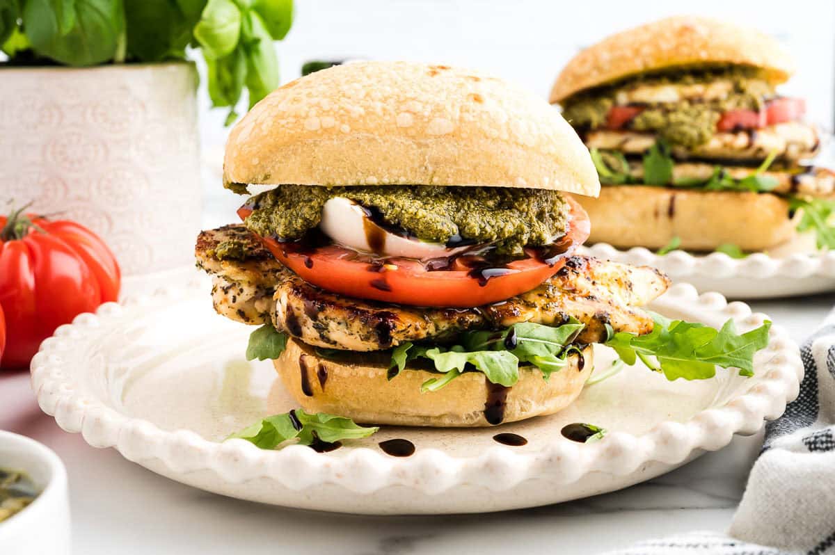 Grilled chicken sandwich with tomato, mozzarella, arugula, pesto, and balsamic glaze on a ciabatta bun, served on a white plate.
