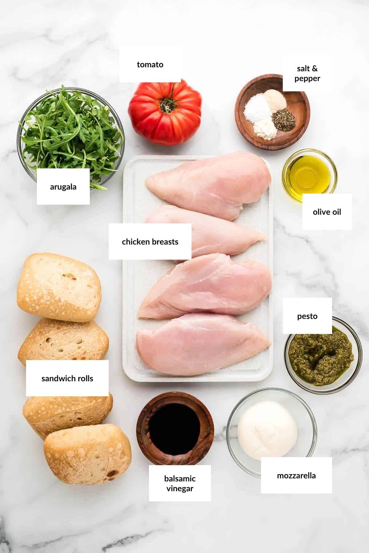 Top-down view of ingredients for a chicken pesto sandwich: arugula, tomato, chicken breasts, sandwich rolls, mozzarella, pesto, olive oil, balsamic vinegar, salt, and pepper, labeled on a white surface.