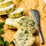 Slices of compound butter with chopped herbs are arranged on brown parchment paper next to a butter knife and fresh parsley.