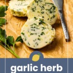 Sliced garlic herb butter on parchment paper with a knife and fresh herbs beside it.
