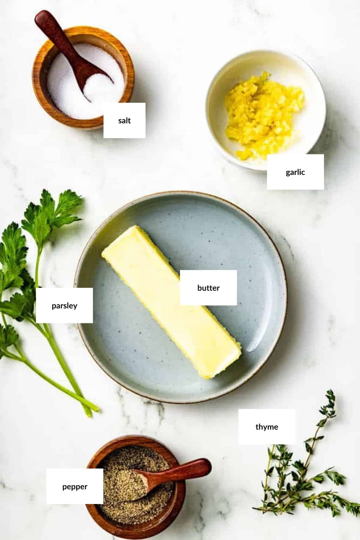 A stick of butter on a plate, surrounded by bowls of salt, minced garlic, pepper, and sprigs of parsley and thyme on a white surface.