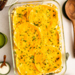 A glass baking dish filled with a layered casserole topped with round tortillas and melted cheese, garnished with chopped herbs. Surrounded by plates, drinks, and condiments on a table.