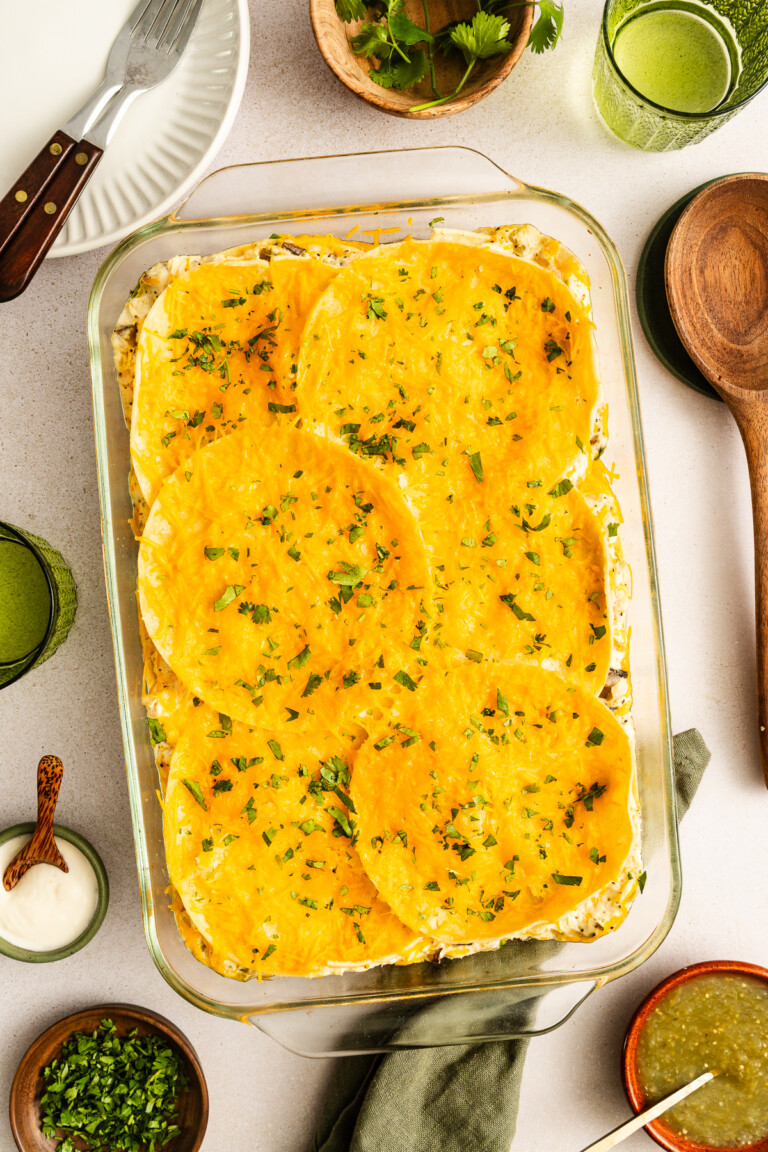 A glass baking dish filled with a layered casserole topped with round tortillas and melted cheese, garnished with chopped herbs. Surrounded by plates, drinks, and condiments on a table.