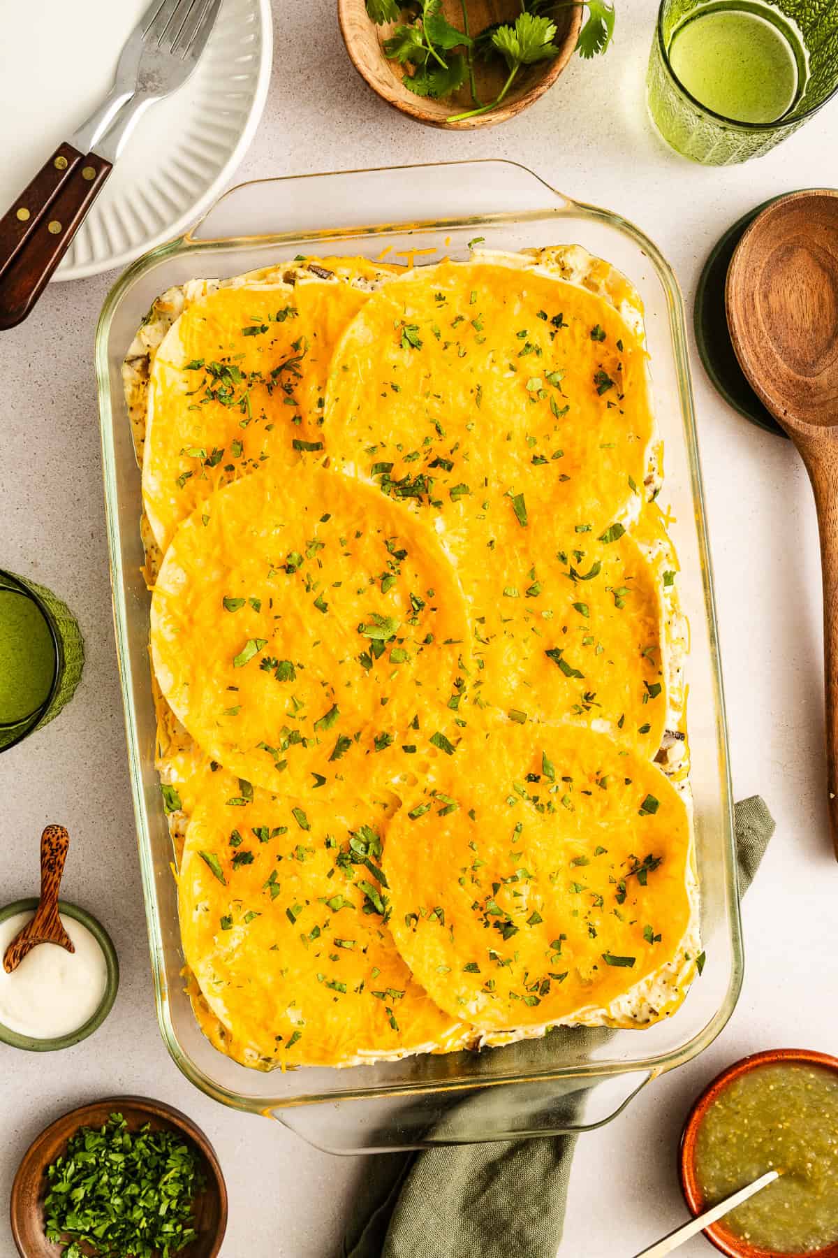 A glass baking dish filled with a layered casserole topped with round tortillas and melted cheese, garnished with chopped herbs. Surrounded by plates, drinks, and condiments on a table.