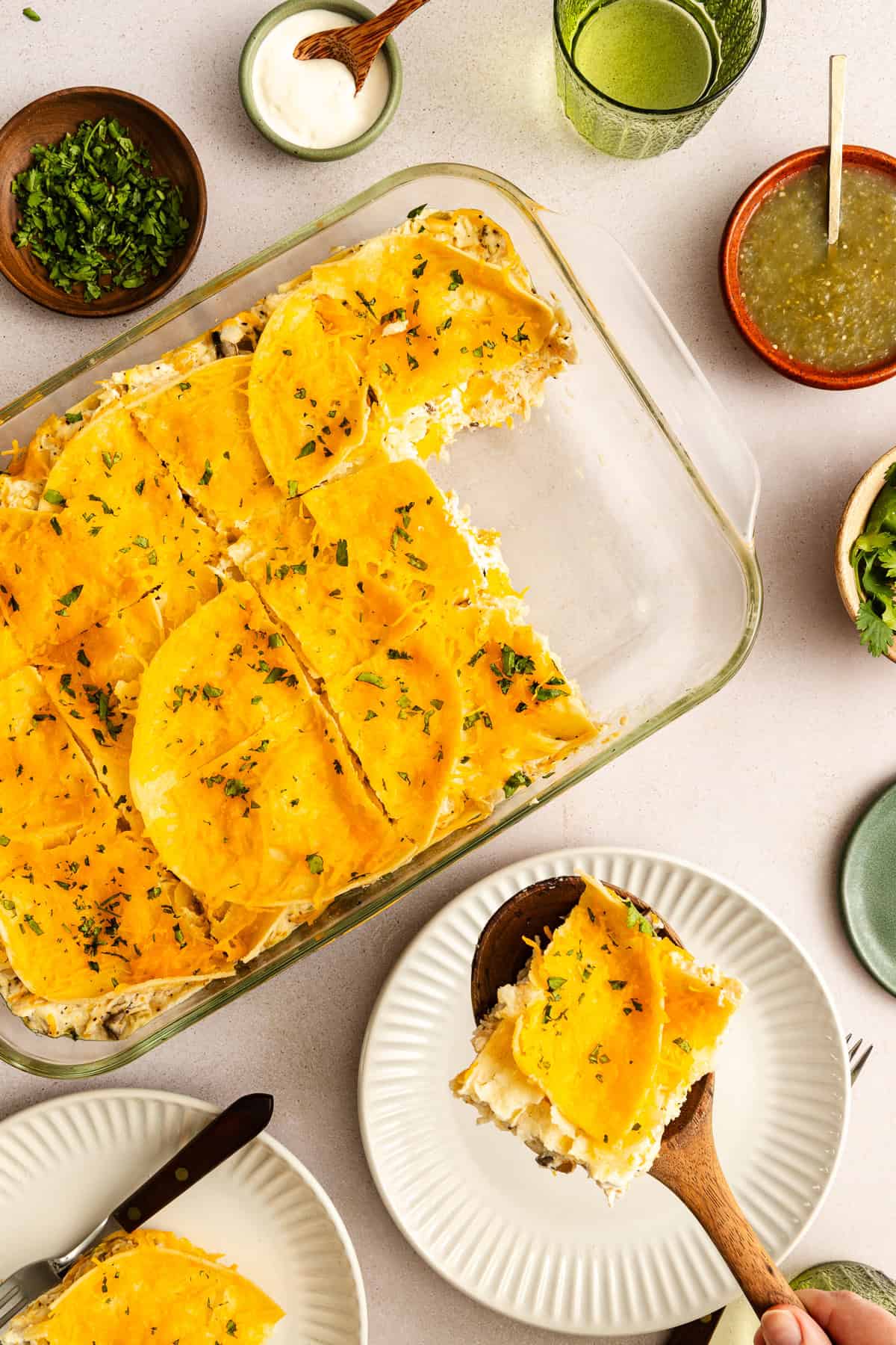 A glass baking dish with sliced, baked casserole topped with yellow tortillas and herbs, served with plates, salsa, sour cream, and cilantro on a light surface.