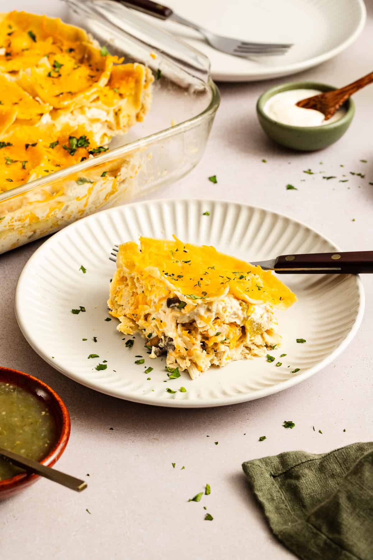 A slice of cheesy casserole on a white plate with a fork, garnished with chopped herbs. The casserole dish and condiments are in the background.