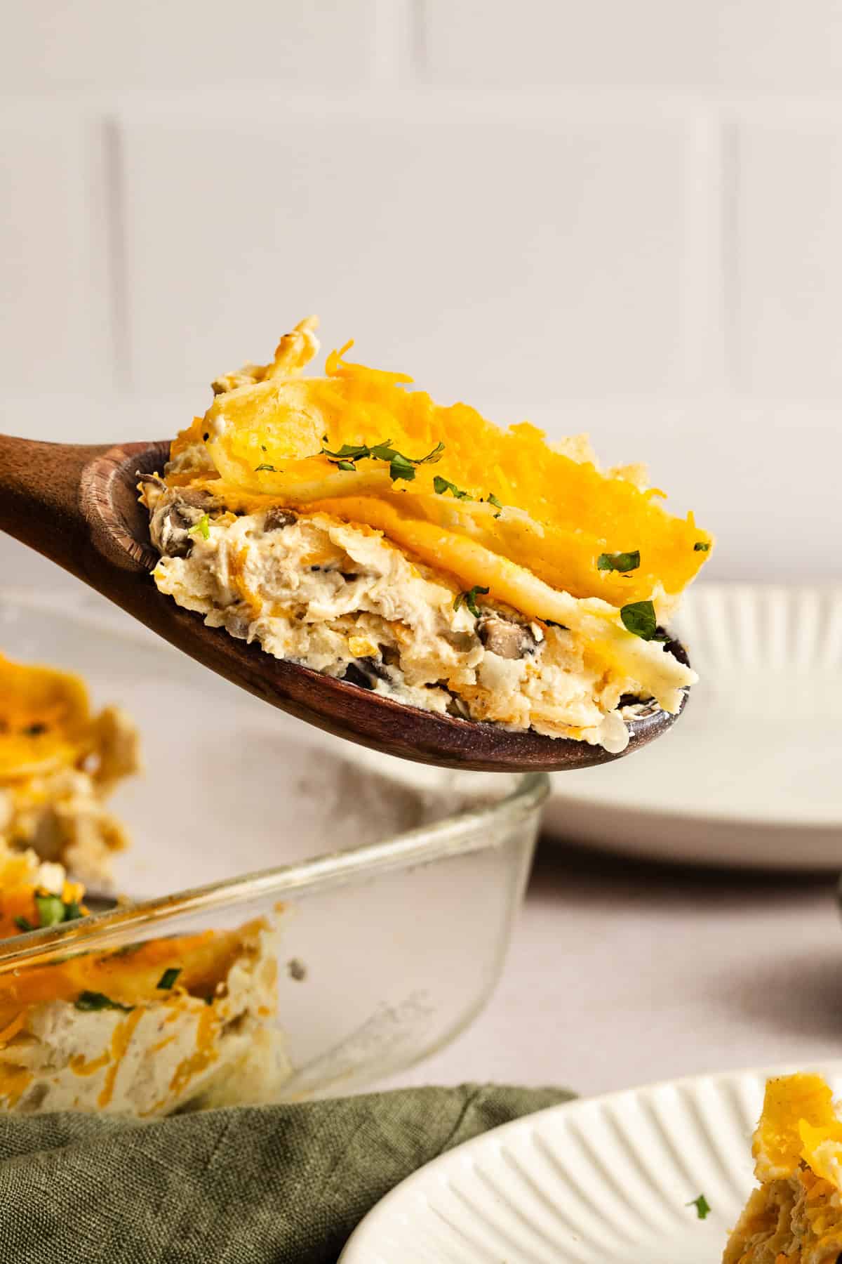 A wooden spoon holds a serving of layered casserole with cheese and herbs above a glass baking dish, with plates and a green napkin nearby.