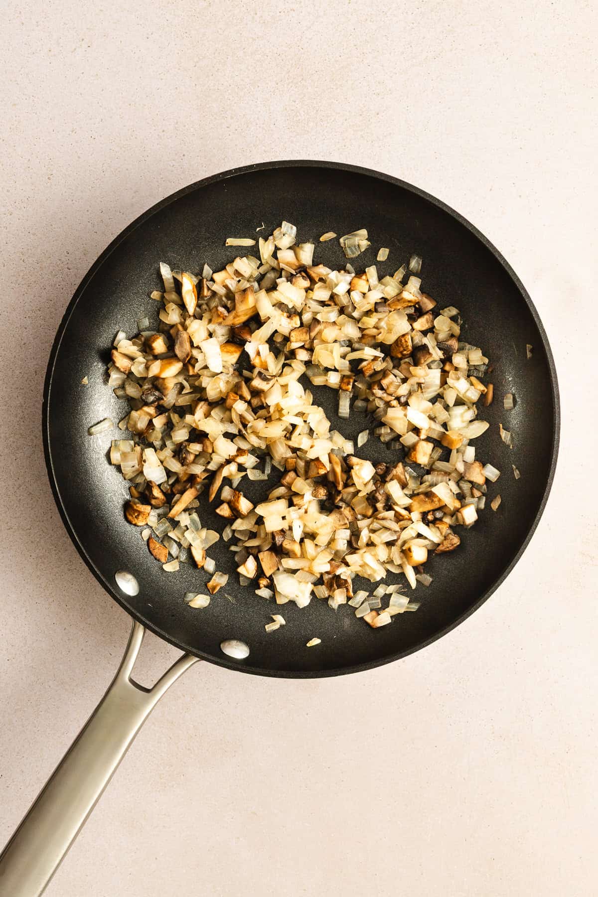 Chopped onions and mushrooms saut&eacute;ing in a black frying pan on a light countertop.